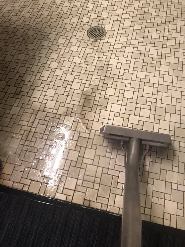 Close-up of a tiled floor being cleaned with a steam cleaner, showcasing the expertise of commercial services in action. A distinct line appears where dirt is removed, unveiling the original tile color.