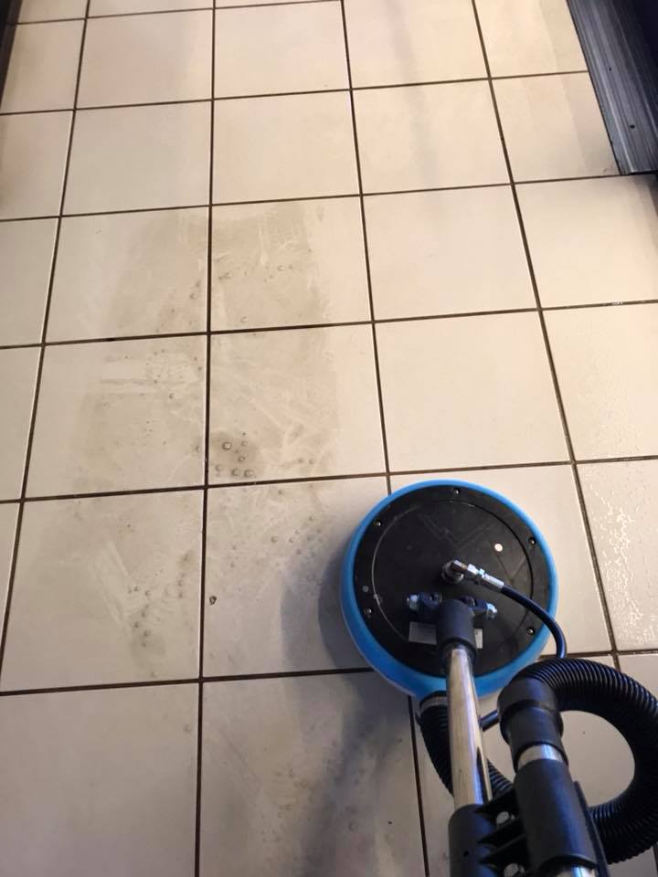 A tile floor is being cleaned with a circular floor cleaning machine, showcasing the expertise of commercial services. The result is a striking line between dirty and clean tiles.