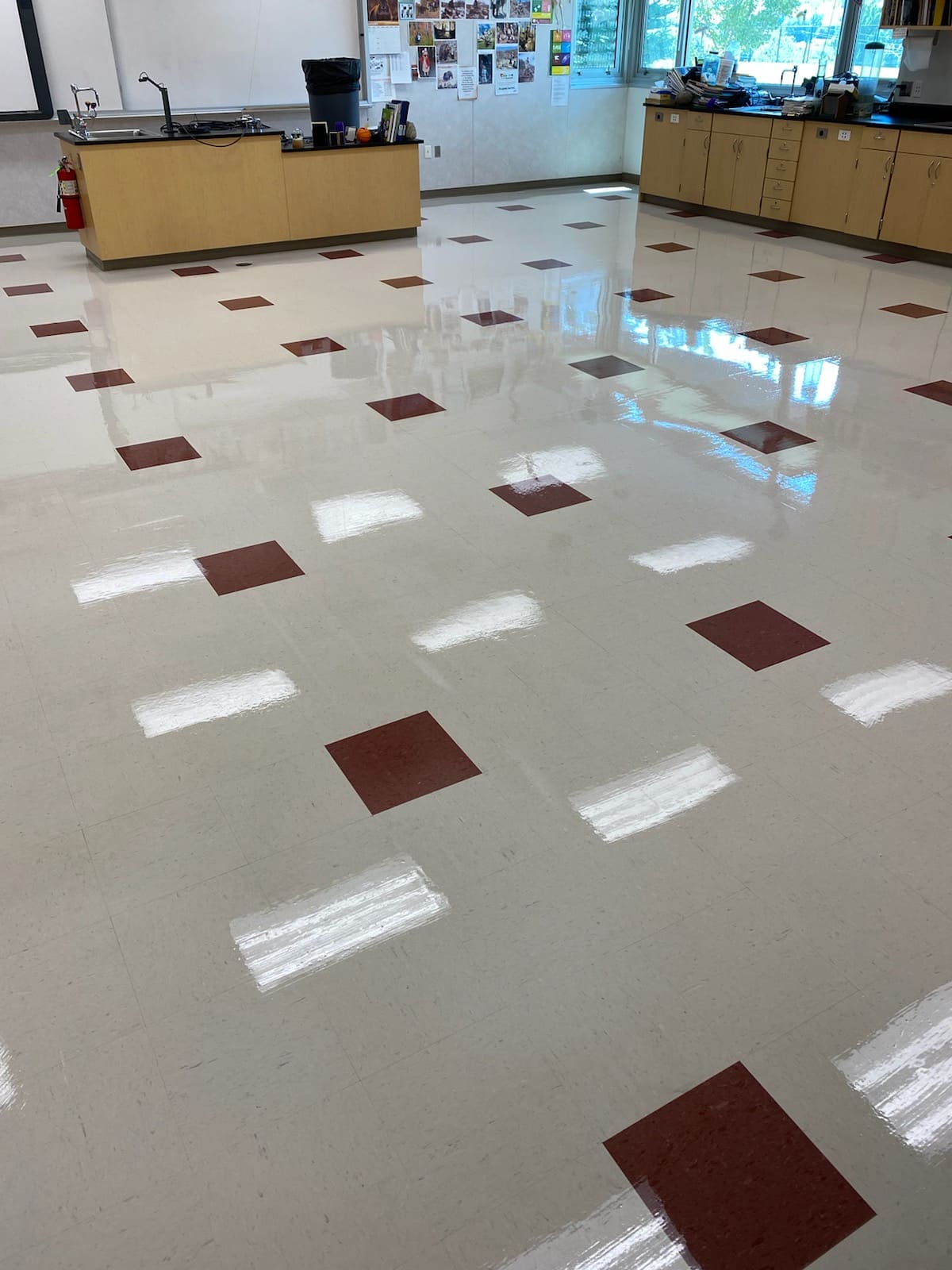 A classroom with a shiny tiled floor, featuring a pattern of large white and smaller red squares, exudes an aura akin to refined commercial services. Wooden cabinets, a counter, and educational posters are visible in the background.